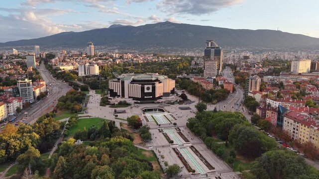 A drone approaches a large cultural complex and public plaza surrounded by city streets with a mountain range in the background.