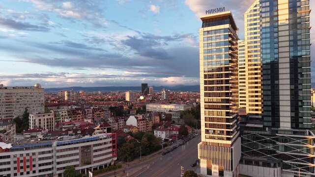 A drone passes modern glass towers and reveals dense urban neighborhoods, a busy boulevard and mountains on the horizon.