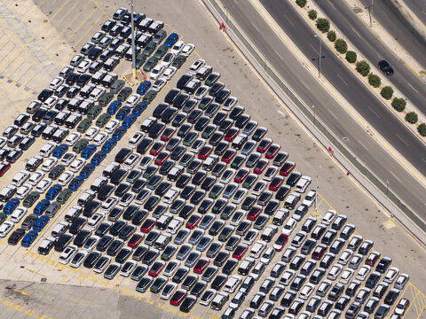 Aerial view of a large parking lot filled with numerous cars arranged in neat rows next to a multi-lane highway in Greece.