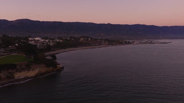 Aerial shot captures the Santa Barbara coastline, beach, and mountains under a twilight sky at dusk in California. Coastal bluffs overlook gentle ocean waves during the evening.
