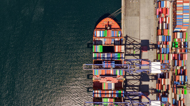 Aerial view of a large container ship being loaded with colorful shipping containers by industrial cranes at a busy port in Greece.