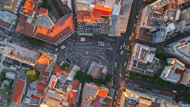 Aerial view of a bustling city square in Greece, featuring intricate stone paving, surrounding historic buildings with terracotta rooftops, and golden hour light casting long shadows.