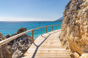 Obraz premium Dhermi beach near the city of Vlore in Albania, Europe. Wooden boardwalk pathway along rocky coastline overlooking the deep blue sea under a clear sky in a scenic coastal location