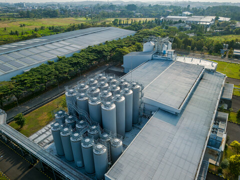 Aerial view of an industrial facility with tall metal storage silos, warehouse buildings with solar panels, and surrounding green landscape in Semarang Regency, Central Java, Indonesia.