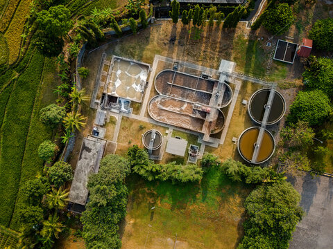 Aerial view of a wastewater treatment plant with circular and oval aeration tanks surrounded by lush green rice terraces and tropical trees in Semarang Regency, Central Java, Indonesia.