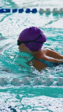 Swimmer in purple cap and goggles moves through pool lanes. Water ripples around focused athlete. Red and blue lane markers define racing path. Intense concentration visible in swimmer's posture