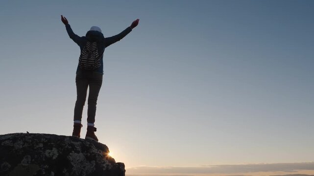 Silhouette of a hiker on a cliff edge raising arms in triumph against a bright sunset.