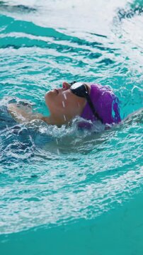 Swimmer in purple cap and goggles glides through clear blue water. Arm extends high, showing focused effort. Head turns slightly, revealing determination. Perfect for fitness, sports