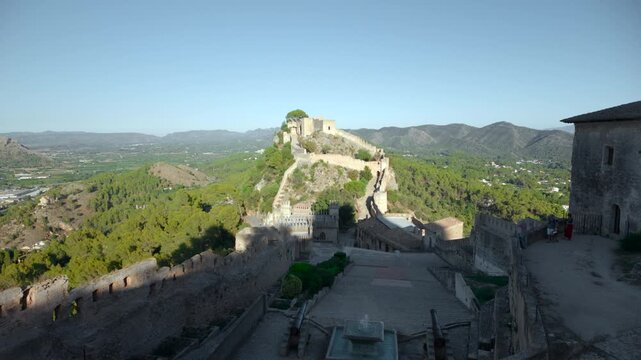 Panoramic view of stone fortress walls on rocky ridge above green valley, distant mountains, terraced walkways, towers, and clear blue sky in bright daylight
