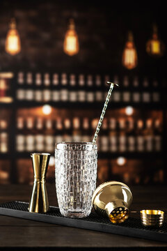 Gold bar tools on black mat. Clear textured glass jigger shaker lid twisted spoon. Blurred bar background.