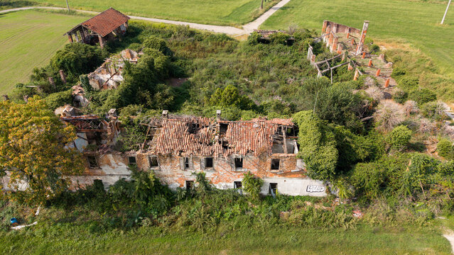 Aerial view of an abandoned farmhouse with crumbling terracotta rooftops and overgrown vegetation surrounded by green fields in Milan, Lombardy, Italy.
