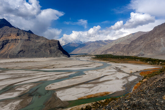 Aerial view of the Shigar Delta, a braided river system with meandering channels and sandy banks set against dramatic mountains under a bright sky, Shigar, Gilgit-Baltistan, Pakistan.