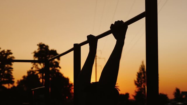 Silhouette of man performing pull ups on horizontal bar during golden sunset workout.