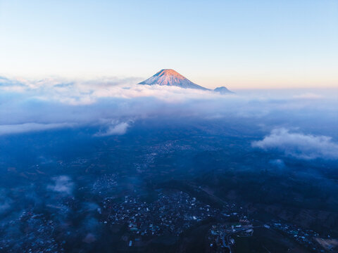 Aerial view of the Dieng Plateau mountain peak rising above a sea of clouds at sunrise, with golden light on the summit and a village visible in the valley below in Indonesia.