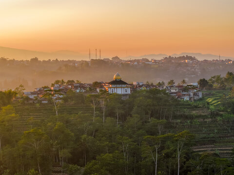 Aerial view of the Dieng Plateau featuring a mosque with a golden dome nestled among lush trees and misty hills under the warm glow of sunrise in Indonesia.