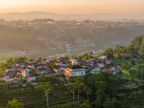 Aerial view of a village with a mosque featuring a golden dome, surrounded by lush terraced fields and misty mountains during golden hour in Indonesia.