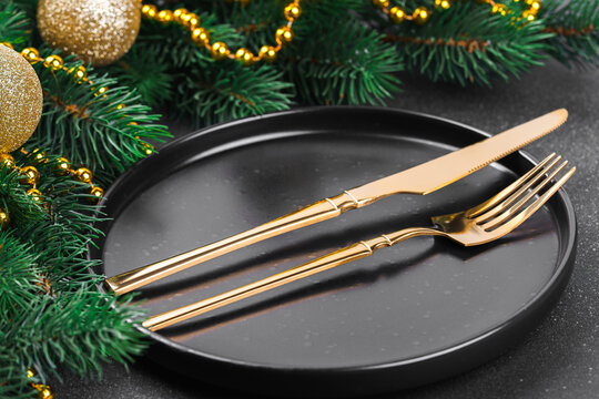 Elegant Christmas table setting with gold cutlery on a black plate, surrounded by evergreen branches, golden ornaments, beads and pinecones on a dark textured background