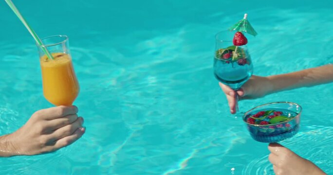 Friends toasting with colorful cocktails in swimming pool during summer vacation.