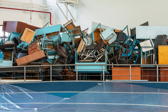 A large and messy heap of old school desks, chairs, and tables stacked chaotically behind a metal railing inside a gymnasium.