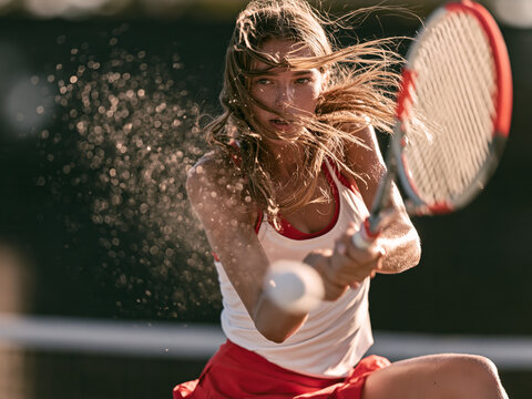 Dynamic action shot of focused female tennis player hitting two-handed backhand swing with racket on outdoor court, athletic competition and sportswear concept.