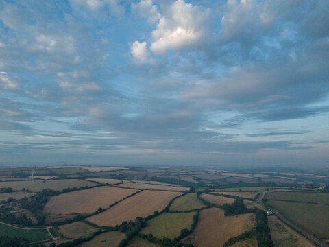 Aerial view of agricultural farmland with harvested fields, hedgerows, and rural tracks under a cloudy sky in Looe, Cornwall, United Kingdom.