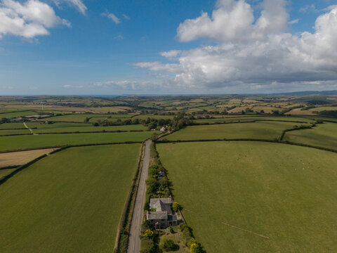 Aerial view of agricultural farmland in Looe featuring green fields, hedgerows, a country road, and scattered farmhouses under a blue sky with clouds. Looe, Cornwall, United Kingdom.