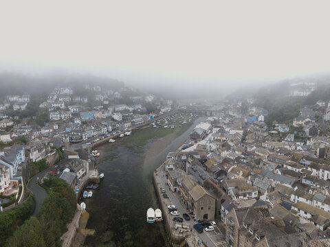 Aerial view of the coastal town of Looe featuring the river estuary, historic buildings, and small boats docked along the banks under morning fog in Looe, Cornwall, United Kingdom.