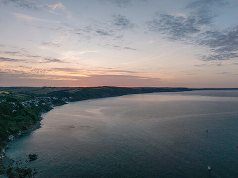 Aerial view of the coastline of Looe at sunrise with calm sea, green cliffs, and soft morning light in Looe, Cornwall, United Kingdom.