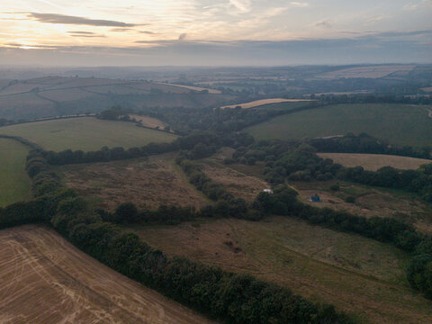 Aerial view of misty rural landscape with agricultural farmland, hedgerows, and rolling hills under a soft sunset sky in Looe, Cornwall, United Kingdom.