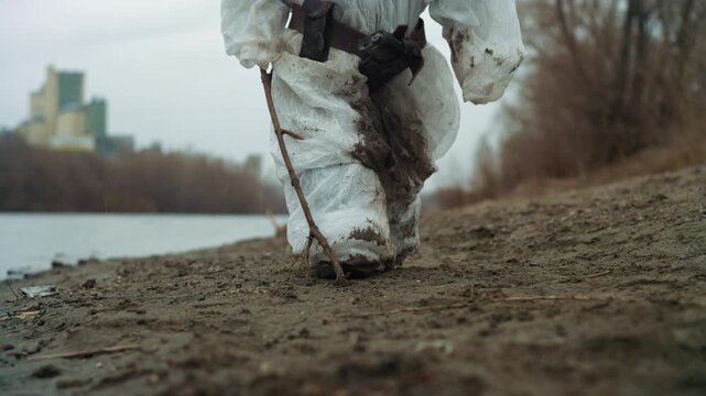 Scavenger unknown closeup boots and staff trudging shore, mud clinging to tattered suit, stick for balance, slow heavy steps, gritty textures of pebble and debris, low angle cinematic detail