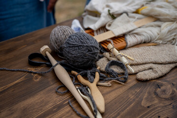 Handmade wool yarn balls, wooden drop spindle, lucet fork, weaving tools, and knitted mittens arranged on rustic wooden table outdoors. © Marina
