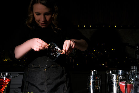 Female bartender decorating martini glass with lime zest - cocktail mixology process. Woman making alcoholic drink at dark bar counter, professional service.