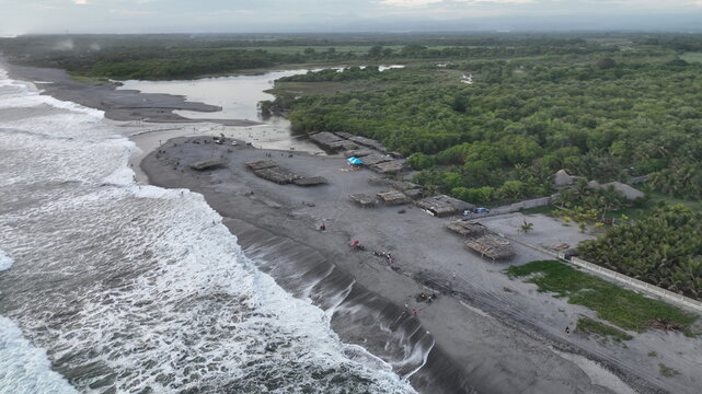 Aerial view of San Marcelino beach with dark volcanic sand, crashing Pacific waves, a coastal lagoon, and thatched-roof huts amidst lush vegetation in La Paz Centro, La Paz, El Salvador.