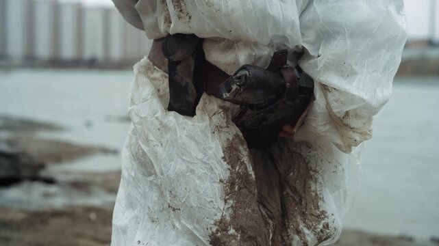Rescue worker cradles injured bird closeup, hands gently holding fragile wet feathers, mudstreaked suit and gloves, field medical kit at waist, tension mixed with tenderness in urban estuary aftermath