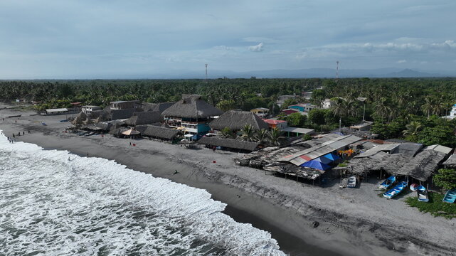 Aerial view of San Marcelino beach featuring thatched-roof structures along the coastline with crashing waves and lush palm trees in La Paz Centro, La Paz, El Salvador.
