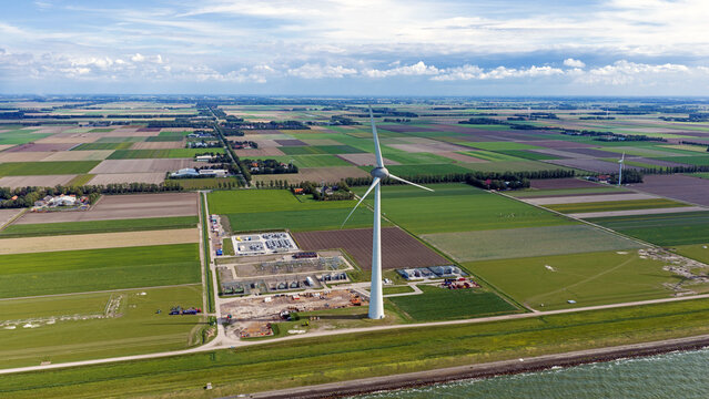 Aerial view of a wind turbine standing in a rural landscape with agricultural fields, a power substation, and a dike near the water in Espel, Flevoland, Netherlands.