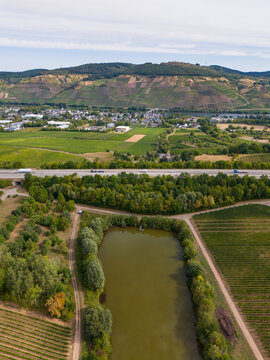 Aerial view of the Moselle valley landscape with lush vineyards, a highway road with vehicles, and a calm pond surrounded by trees in Trier-Saarburg, Rhineland-Palatinate, Germany.