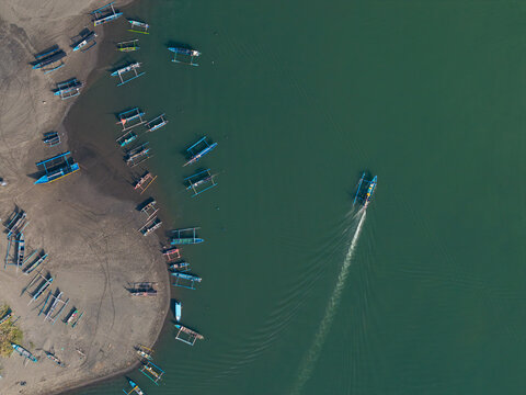 Aerial view of Pengambengan National Fishing Port with numerous traditional fishing boats moored along the sandy shoreline and a boat leaving a wake in Kecamatan Jembrana, Bali, Indonesia.