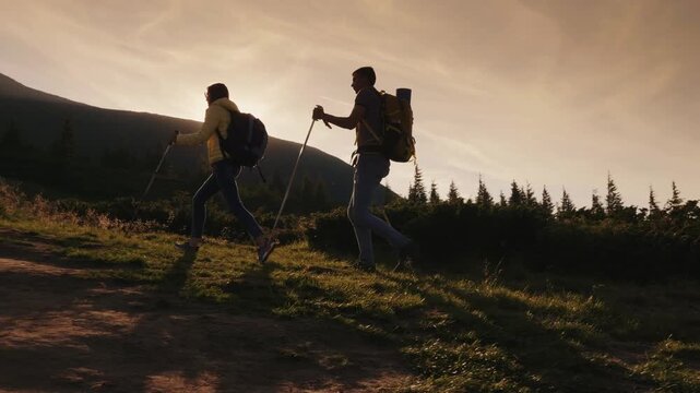 Dynamic cinematic footage of two hikers with backpacks and trekking poles ascending a mountain path at sunset, sun's rays bursting from behind them, highlighting their silhouettes as they move through