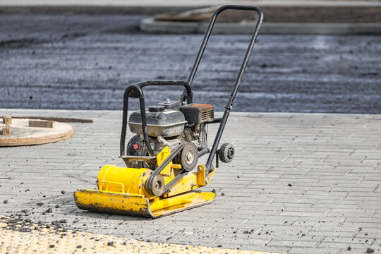 A vibratory plate for compacting asphalt and soil is located on a paved area where new asphalt has been laid. The equipment is currently not in use