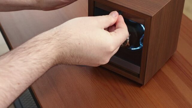 A hand placing a luxury watch into an automatic wooden winder box on a dark brown desk, representing precision maintenance, horology care, and high-end lifestyle product organization