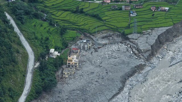 Aerial view of a landslide area with debris near a river, surrounded by green terraced rice fields and a winding road in Betrawoti Bazaar, Bagmati Province, Nepal.