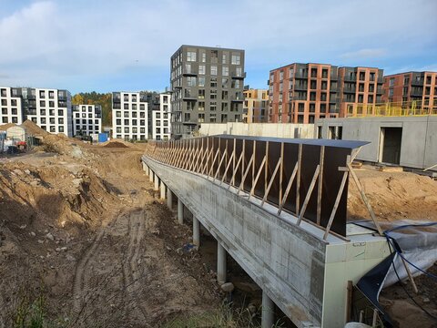 A construction site featuring a partially completed elevated concrete walkway or bridge structure supported by cylindrical pillars, with wooden bracing and dark formwork panels visible on top. The