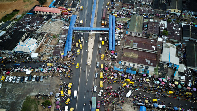 Aerial view of the busy Agege intersection with yellow buses, crowded market stalls, and pedestrians moving around the blue pedestrian bridge in Lagos, Lagos, Nigeria.