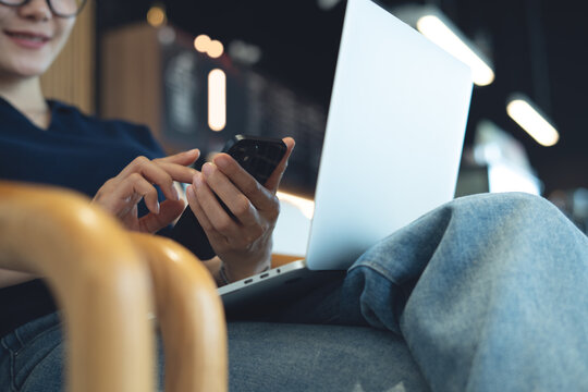 Asian woman smiling during using smartphone for online messaging with laptop on her lap, remote at cafe, digital nomad lifestyle