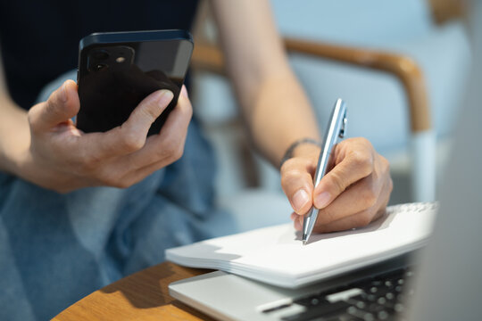 Business woman using smartphone searching and browsing the internet, writing in notebook with laptop on table, remote working