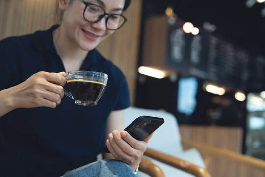 Smiling woman hand holding a cup of black coffee and using smartphone at cafe, modern people lifestyle