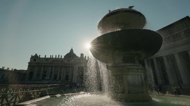 Sunlight rays shine through fountain water droplets on saint peters square with saint peters basilica in background on sunny day.