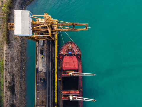 Aerial view of a cargo ship docked at a port with a large yellow crane, turquoise water, and industrial infrastructure in Cilacap, Central Java, Indonesia.