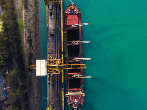 Aerial view of a large cargo ship docked at a port terminal with yellow cranes and turquoise water in Cilacap, Central Java, Indonesia.